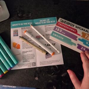 August SketchBox unboxing featuring professional alcohol markers, Copic Sketch marker, Sakura Pigma Brush Pen, Faber-Castell Polychromos pencil, and SketchBox Signature marker paper before creating the monthly project.